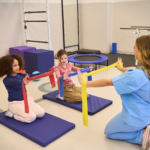 A pediatric therapist in scrubs working with two young children using resistance bands in a colorful therapy gym.