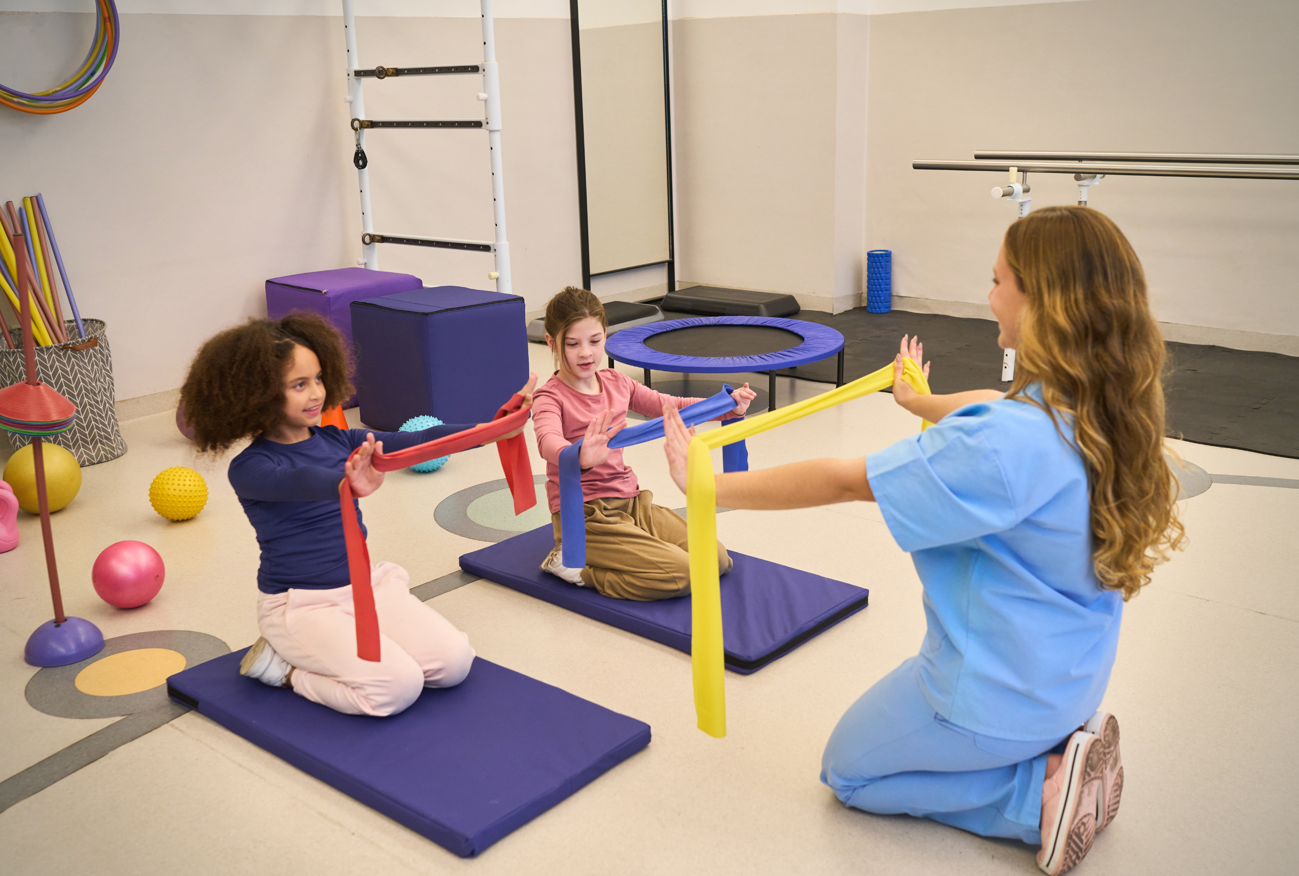 A pediatric therapist in scrubs working with two young children using resistance bands in a colorful therapy gym.