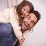 A father laughing and giving his young daughter a piggyback ride at home, representing dad involvement in children's therapy support.