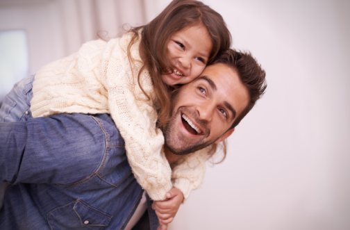 A father laughing and giving his young daughter a piggyback ride at home, representing dad involvement in children's therapy support.