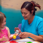 A registered behavior technician in scrubs working one-on-one with a young girl using animal toys during ABA therapy.
