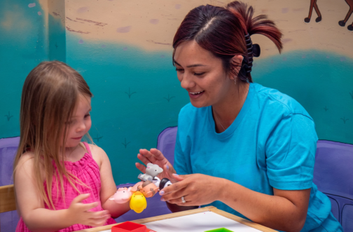 A registered behavior technician in scrubs working one-on-one with a young girl using animal toys during ABA therapy.
