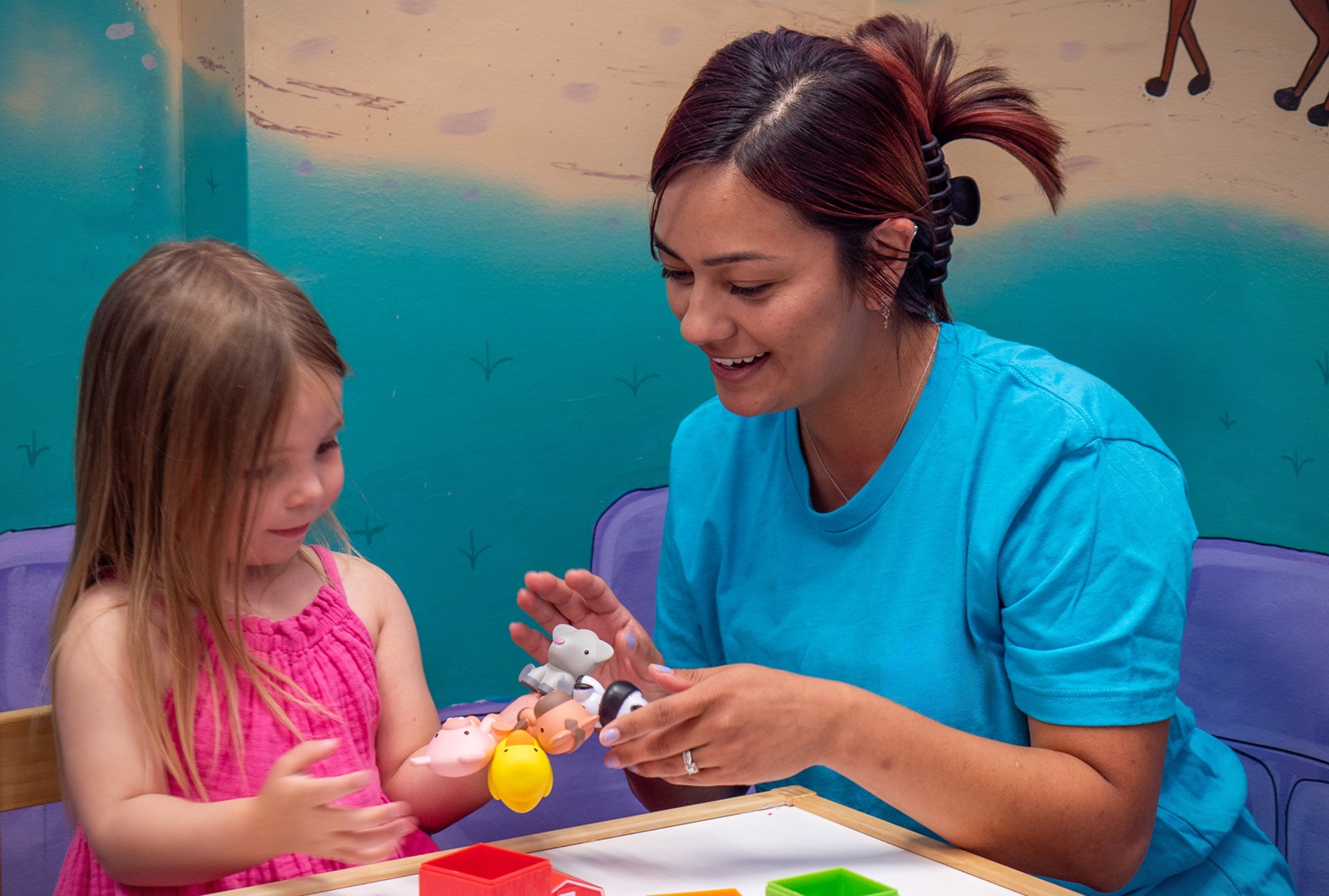 A registered behavior technician in scrubs working one-on-one with a young girl using animal toys during ABA therapy.