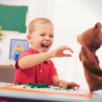 A delighted toddler reaching toward a hand puppet during a play-based ABA therapy session in a bright classroom setting.