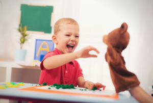 A delighted toddler reaching toward a hand puppet during a play-based ABA therapy session in a bright classroom setting.