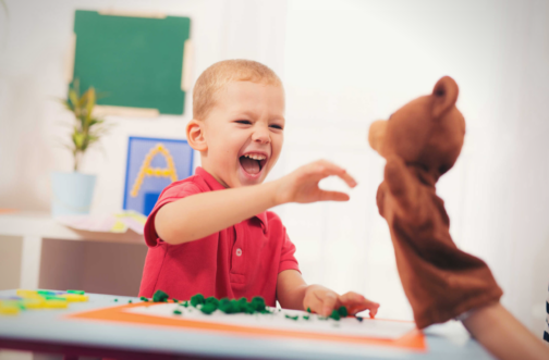 A delighted toddler reaching toward a hand puppet during a play-based ABA therapy session in a bright classroom setting.