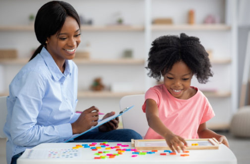 A BCBA therapist taking notes while a young girl works on a learning activity during an ABA therapy session.