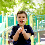 A young boy with autism smiling and playing outdoors at a playground during summer.