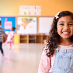 A young girl smiling confidently in a preschool classroom, representing school readiness skills built through ABA therapy for children with autism.