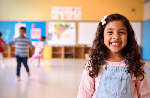 A young girl smiling confidently in a preschool classroom, representing school readiness skills built through ABA therapy for children with autism.