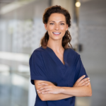 Smiling female clinician in navy scrubs standing confidently with arms crossed in a clinic hallway