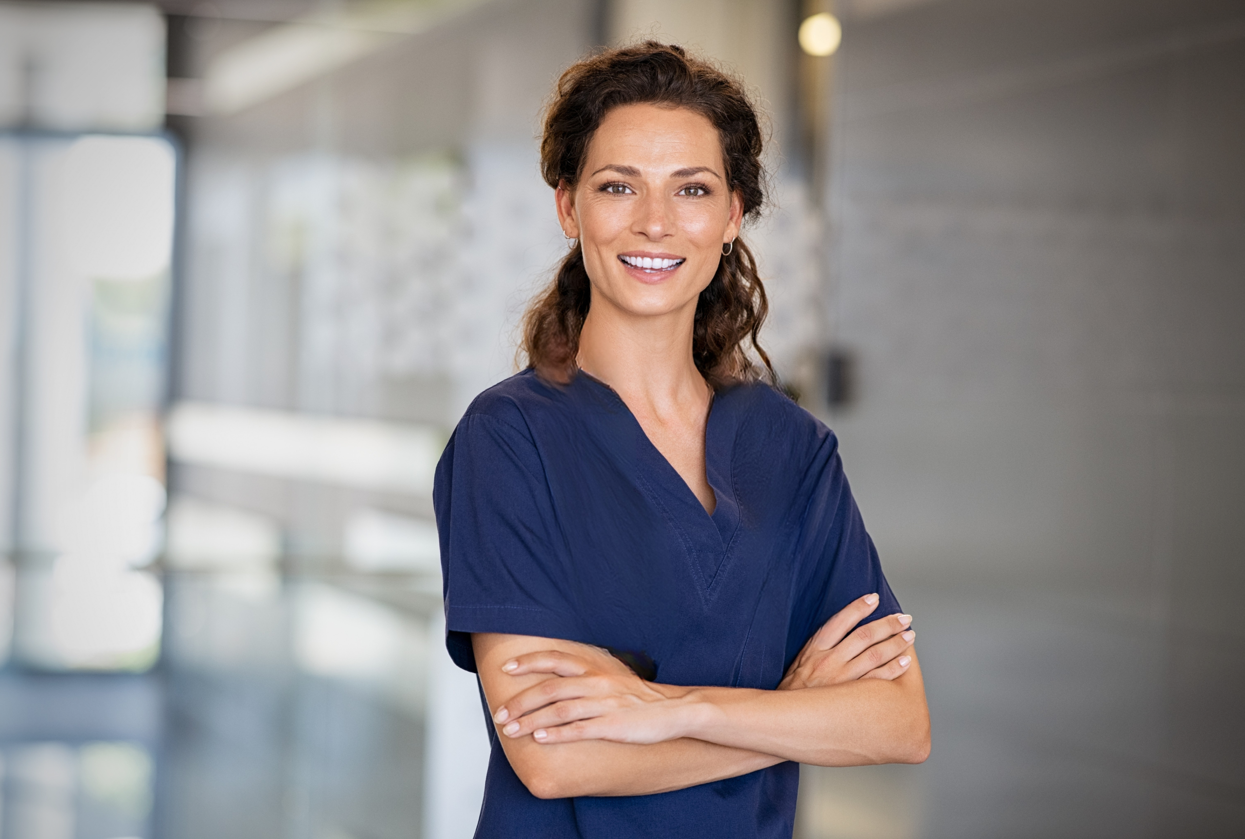 Smiling female clinician in navy scrubs standing confidently with arms crossed in a clinic hallway