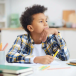 Thoughtful young boy resting his chin on his hand while working at a desk with school supplies