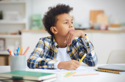 Thoughtful young boy resting his chin on his hand while working at a desk with school supplies