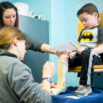 A physical therapist fitting a young boy in a Batman shirt with a serial cast on his lower leg while another therapist assists during a pediatric physical therapy session.