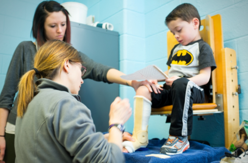 A physical therapist fitting a young boy in a Batman shirt with a serial cast on his lower leg while another therapist assists during a pediatric physical therapy session.