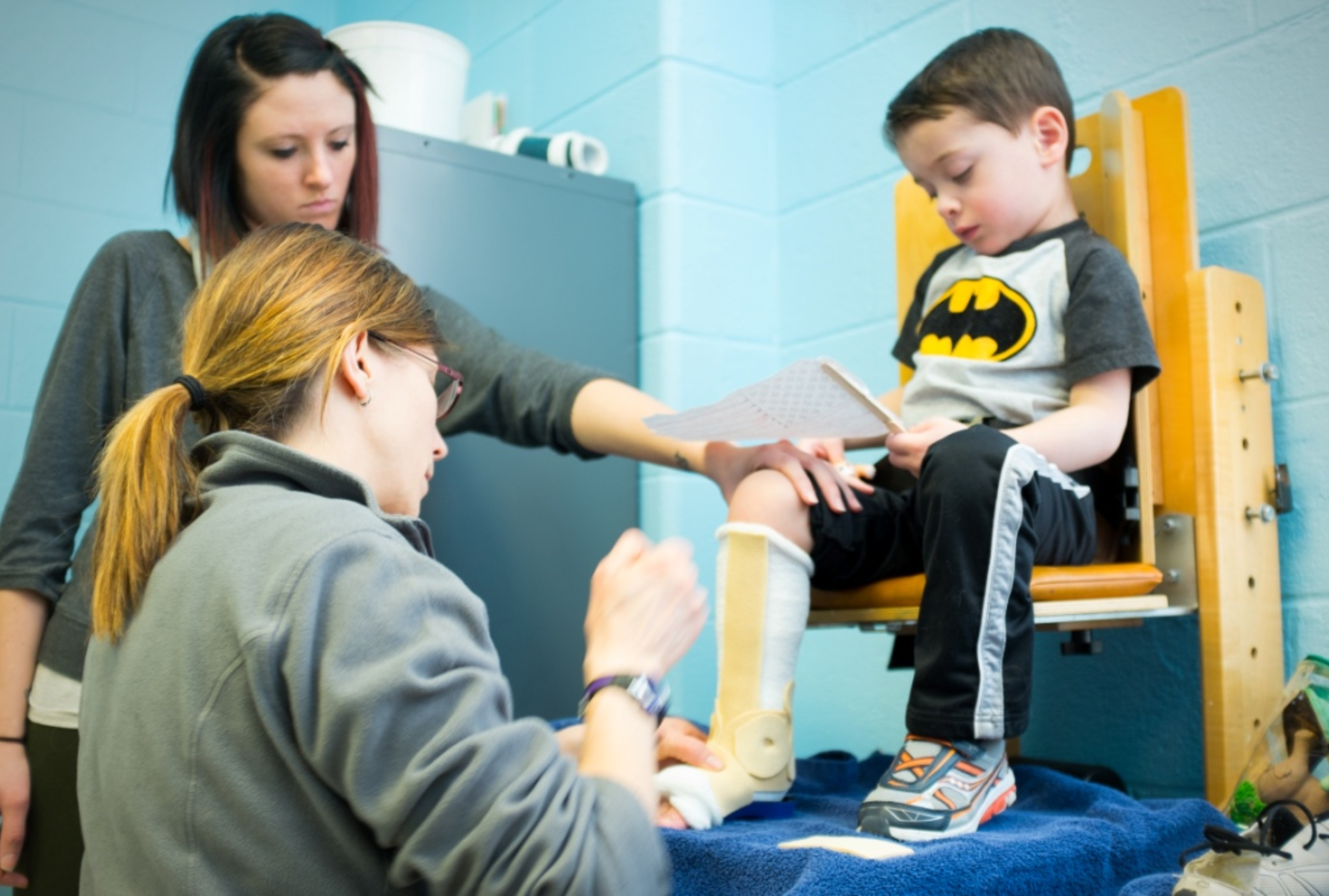 A physical therapist fitting a young boy in a Batman shirt with a serial cast on his lower leg while another therapist assists during a pediatric physical therapy session.