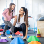 A pediatric physical therapist smiling and holding hands with a young girl wearing an ankle-foot orthosis as she steps across colorful balance blocks in a therapy gym.