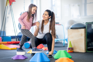 A pediatric physical therapist smiling and holding hands with a young girl wearing an ankle-foot orthosis as she steps across colorful balance blocks in a therapy gym.