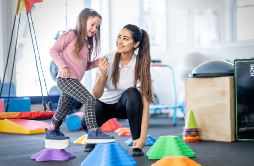 A pediatric physical therapist smiling and holding hands with a young girl wearing an ankle-foot orthosis as she steps across colorful balance blocks in a therapy gym.