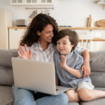Mother and son waving at a laptop screen during a virtual autism evaluation at home