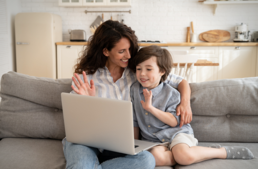 Mother and son waving at a laptop screen during a virtual autism evaluation at home