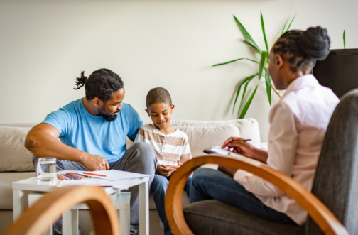 A father and young son sit together on a couch talking with a pediatric counselor taking notes during a family counseling session.