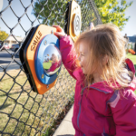 A young girl in a pink jacket smiling while playing with an interactive mirror panel on a playground's chain-link fence, representing the safety fenced-in playgrounds offer families.