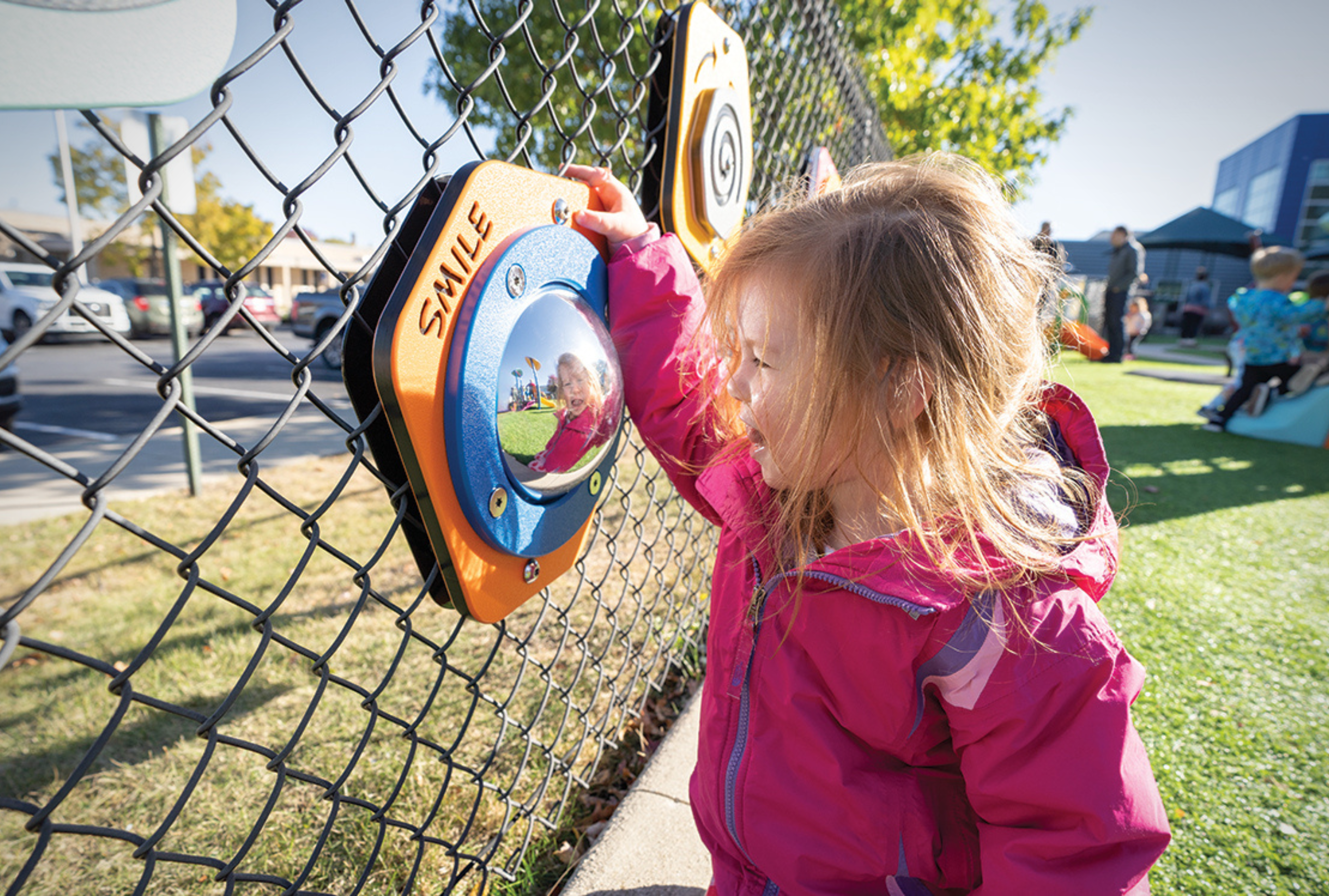 A young girl in a pink jacket smiling while playing with an interactive mirror panel on a playground's chain-link fence, representing the safety fenced-in playgrounds offer families.