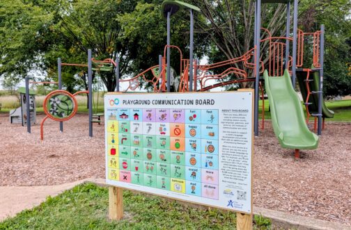 A colorful AAC playground communication board with picture symbols for words like play, swing, help, and all done stands at the entrance to a Mokena park playground, donated by Westside Children's Therapy.