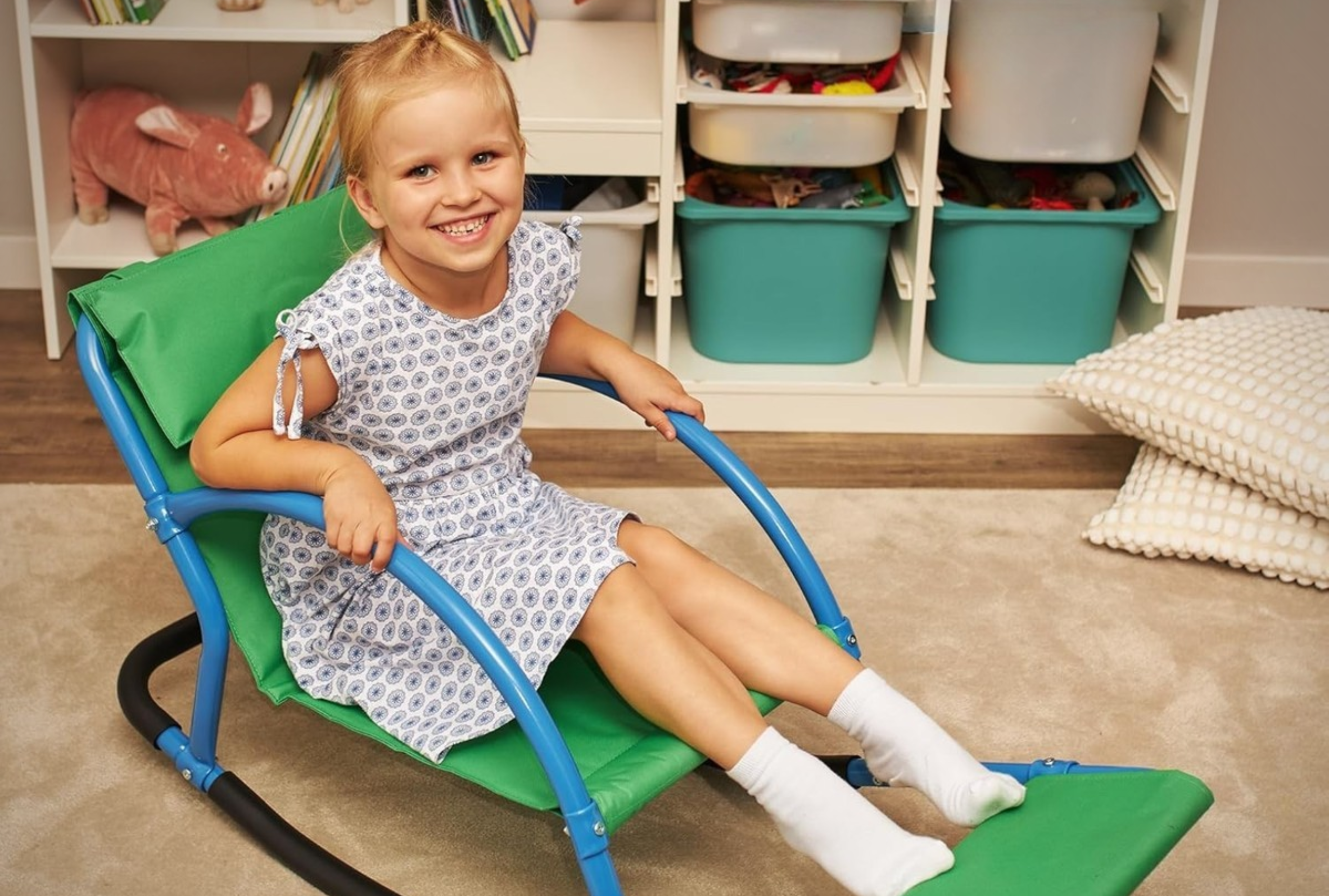 A young girl smiles while sitting in a green and blue rocking chair with a footrest, holding onto the armrests in a playroom.