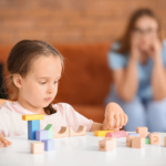 A young girl plays intently with colorful building blocks at a table while her mother watches from a distance with a concerned expression.