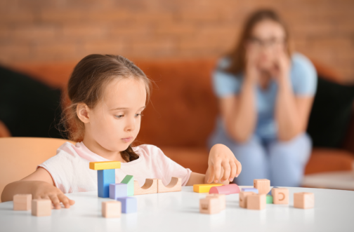 A young girl plays intently with colorful building blocks at a table while her mother watches from a distance with a concerned expression.