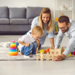 A smiling mother and father play on the floor with their young son, surrounded by stacking rings, blocks, and wooden figures.