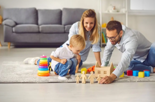 A smiling mother and father play on the floor with their young son, surrounded by stacking rings, blocks, and wooden figures.
