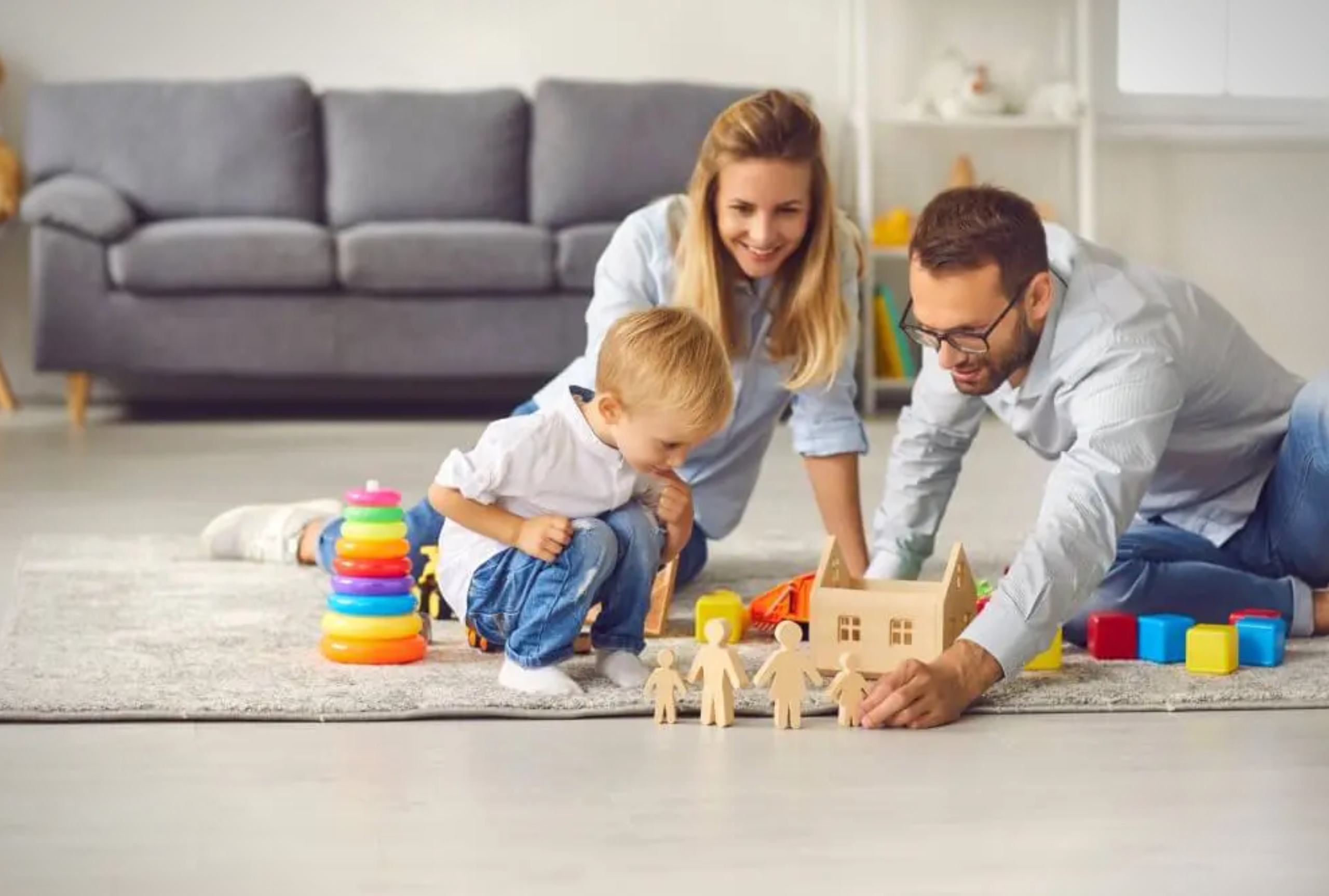 A smiling mother and father play on the floor with their young son, surrounded by stacking rings, blocks, and wooden figures.