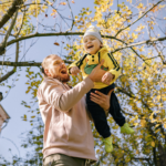 A father laughs while lifting his smiling young son into the air under golden fall leaves, celebrating a joyful outdoor moment together.