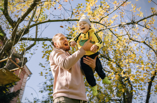 A father laughs while lifting his smiling young son into the air under golden fall leaves, celebrating a joyful outdoor moment together.