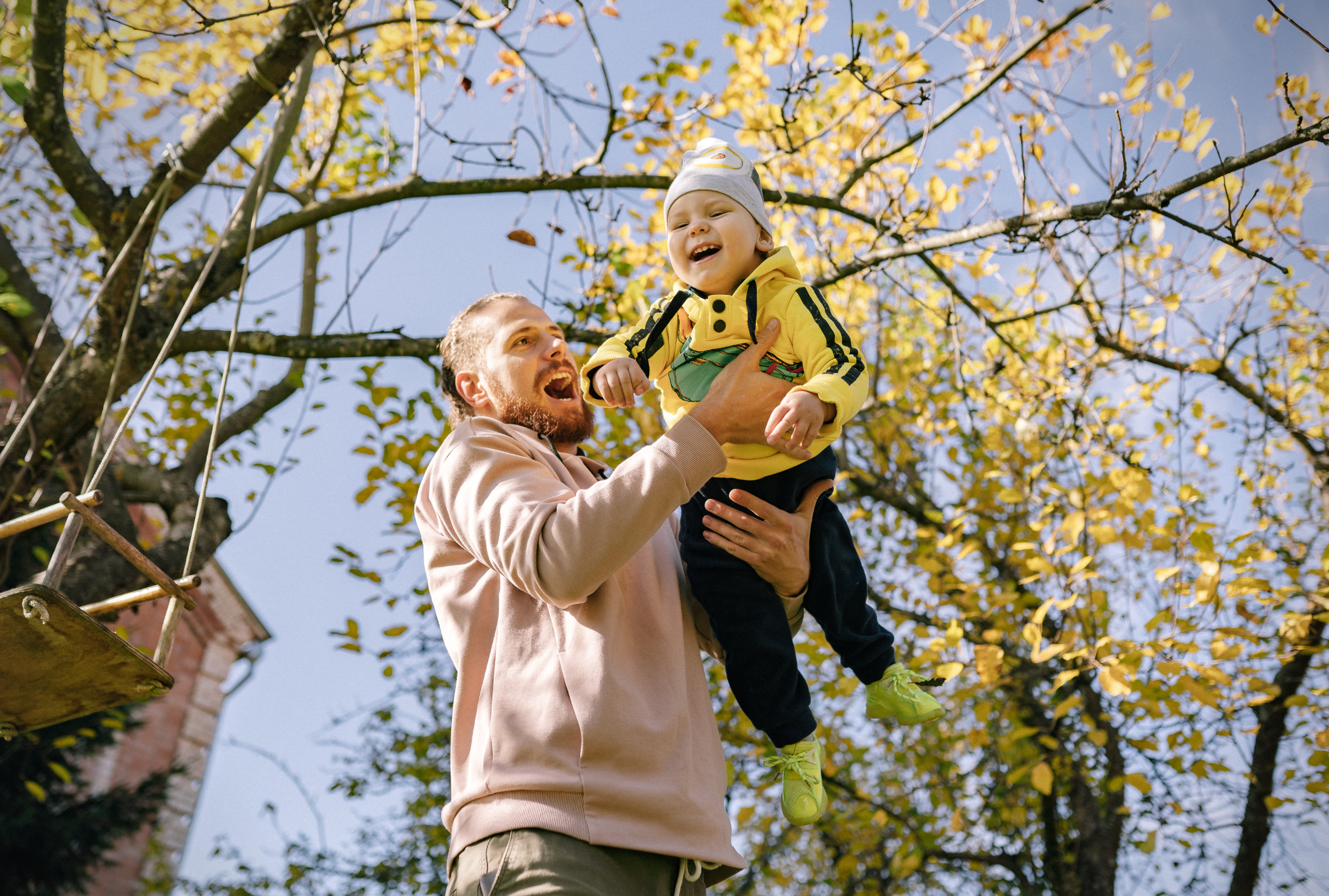 A father laughs while lifting his smiling young son into the air under golden fall leaves, celebrating a joyful outdoor moment together.
