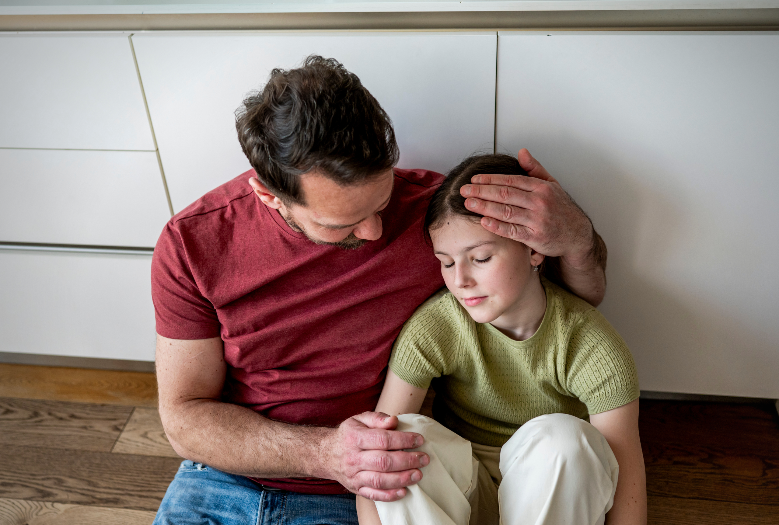 A father gently comforts his young daughter, resting his hand on her head as she leans into him during a quiet moment at home.