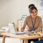 A smiling ABA clinician takes notes on a clipboard beside a laptop while a young boy works on a colorful peg activity at a therapy table.