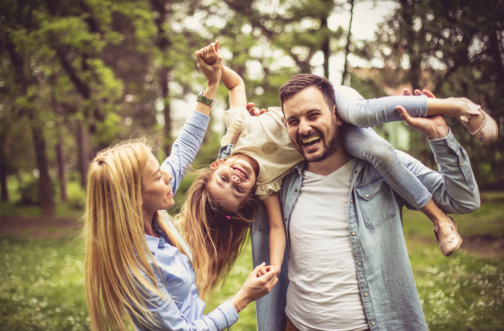 A laughing father carries his giggling daughter across his shoulders as her mother holds her hand in a sunny park.