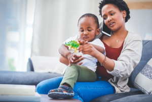 A mother on the phone holds her toddler on her lap as he plays with a toy dinosaur, representing a parent staying proactive while on a waitlist for pediatric therapy.