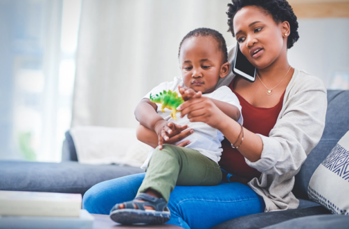 A mother on the phone holds her toddler on her lap as he plays with a toy dinosaur, representing a parent staying proactive while on a waitlist for pediatric therapy.