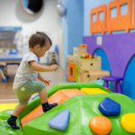 A toddler in socks carefully climbs over a colorful padded play structure at an indoor children's gym.