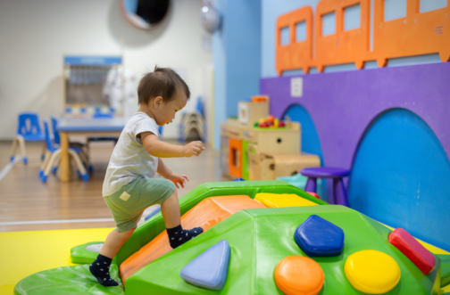 A toddler in socks carefully climbs over a colorful padded play structure at an indoor children's gym.