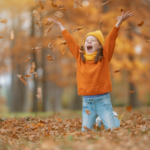 A joyful young girl in an orange sweater and yellow hat kneels in a pile of fall leaves and tosses them into the air, representing seasonal activities that build strength, balance, and coordination.