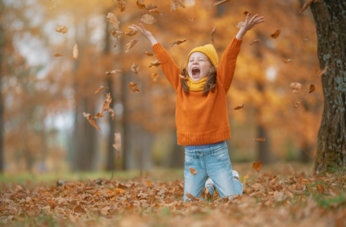 A joyful young girl in an orange sweater and yellow hat kneels in a pile of fall leaves and tosses them into the air, representing seasonal activities that build strength, balance, and coordination.