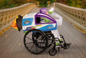 A child in a space ranger costume cheers with a raised fist while seated in a wheelchair transformed into a spaceship with a costume cover, representing adaptive Halloween costume ideas for kids who use wheelchairs.