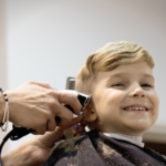 A smiling young boy sits in a salon chair wearing a cape while a stylist trims his hair with clippers, representing strategies that help a child with autism handle haircuts.